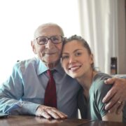 Positive senior man in formal wear and eyeglasses hugging with young lady while sitting at table